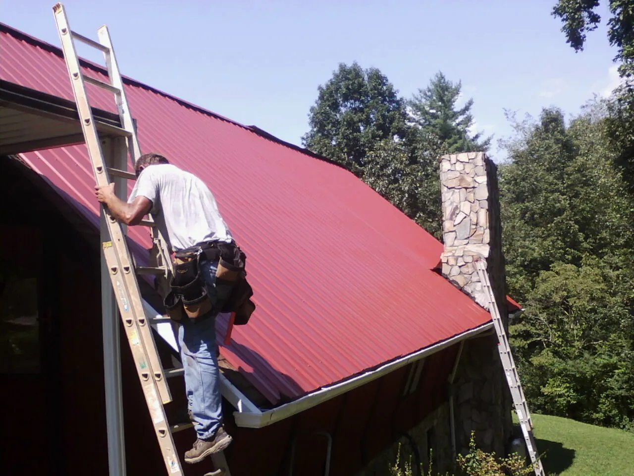 Roofer on a ladder beside a red metal roof and stone chimney — Soffit & Fascia Repair by Roofing Brothers in Powell Butte